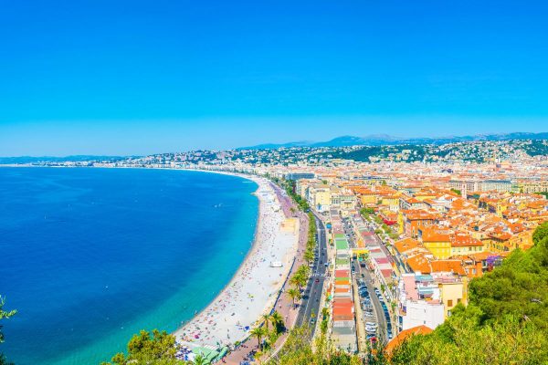 Coastal city with a curved beach, blue sea, and orange-roofed buildings along a seaside promenade.