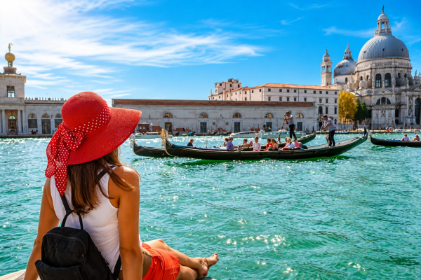 Woman with a bright red sun hat and backpack sits at the edge of a turquoise canal, watching gondolas pass by with a grand cathedral in the background (Venice).